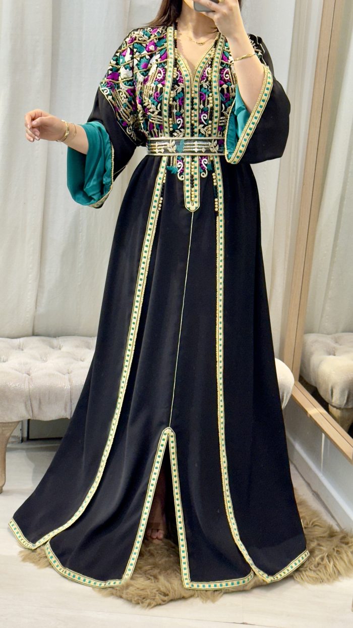 Femme portant un caftan marocain noir avec broderies dorées et roses, ceinture à la taille, manches larges et doublure satinée verte, debout dans une cabine d’essayage.
