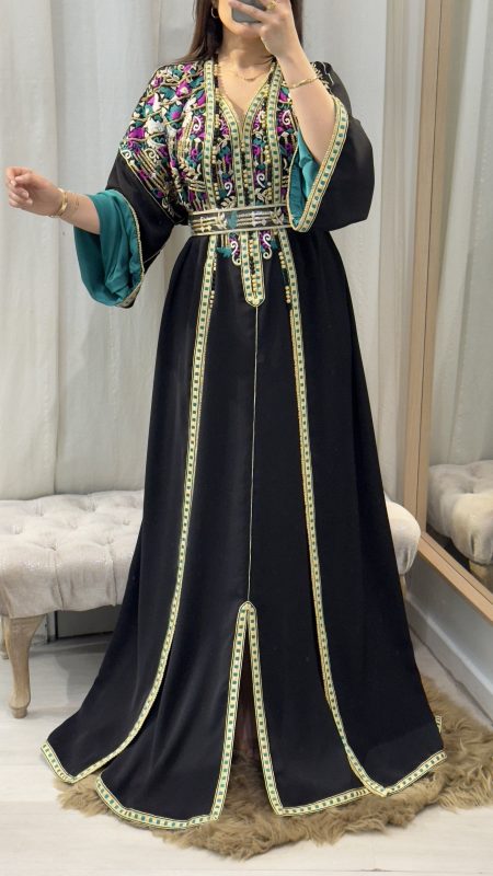 accueil 6 Femme portant un caftan marocain noir avec broderies dorées et roses, ceinture à la taille, manches larges et doublure satinée verte, debout dans une cabine d’essayage.