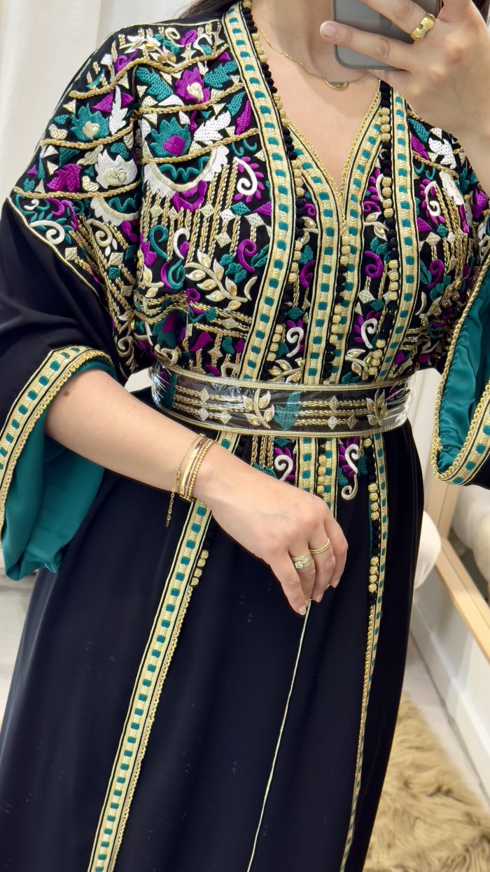 Femme portant un caftan marocain noir avec broderies dorées et roses, ceinture à la taille, manches larges et doublure satinée verte, debout dans une cabine d’essayage.