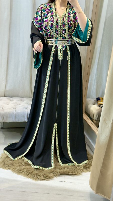 Femme portant un caftan marocain noir avec broderies dorées et roses, ceinture à la taille, manches larges et doublure satinée verte, debout dans une cabine d’essayage.