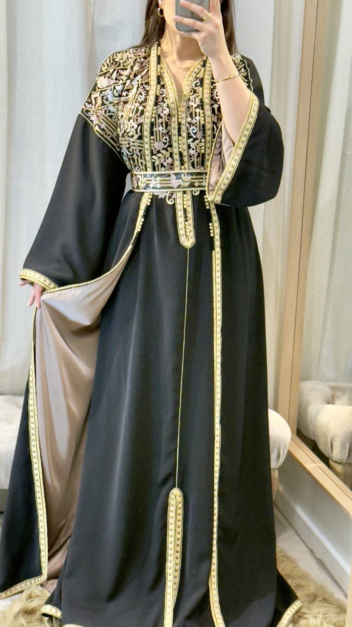 Femme portant un caftan marocain noir avec broderies dorées et roses, ceinture à la taille, manches larges et doublure satinée beige, debout dans une cabine d’essayage.