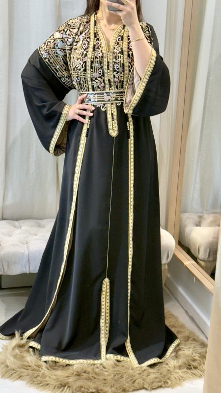 accueil 8 Femme portant un caftan marocain noir avec broderies dorées et roses, ceinture à la taille, manches larges et doublure satinée beige, debout dans une cabine d’essayage.