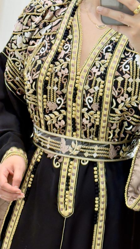 Femme portant un caftan marocain noir avec broderies dorées et roses, ceinture à la taille, manches larges et doublure satinée beige, debout dans une cabine d’essayage.