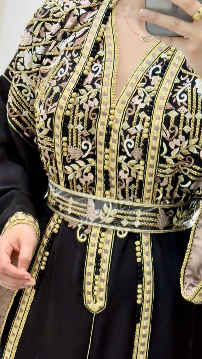 Femme portant un caftan marocain noir avec broderies dorées et roses, ceinture à la taille, manches larges et doublure satinée beige, debout dans une cabine d’essayage.
