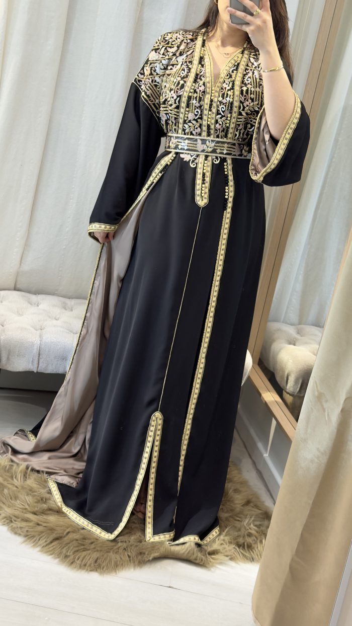 Femme portant un caftan marocain noir avec broderies dorées et roses, ceinture à la taille, manches larges et doublure satinée beige, debout dans une cabine d’essayage.