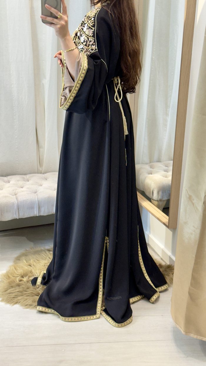 Femme portant un caftan marocain noir avec broderies dorées et roses, ceinture à la taille, manches larges et doublure satinée beige, debout dans une cabine d’essayage.