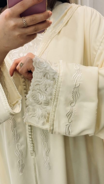 “Djellaba Kmiss femme pour Omra, tissu léger et fluide, broderies larges au buste et aux bouts de manches, couleur écru, coupe ample et élégante.”